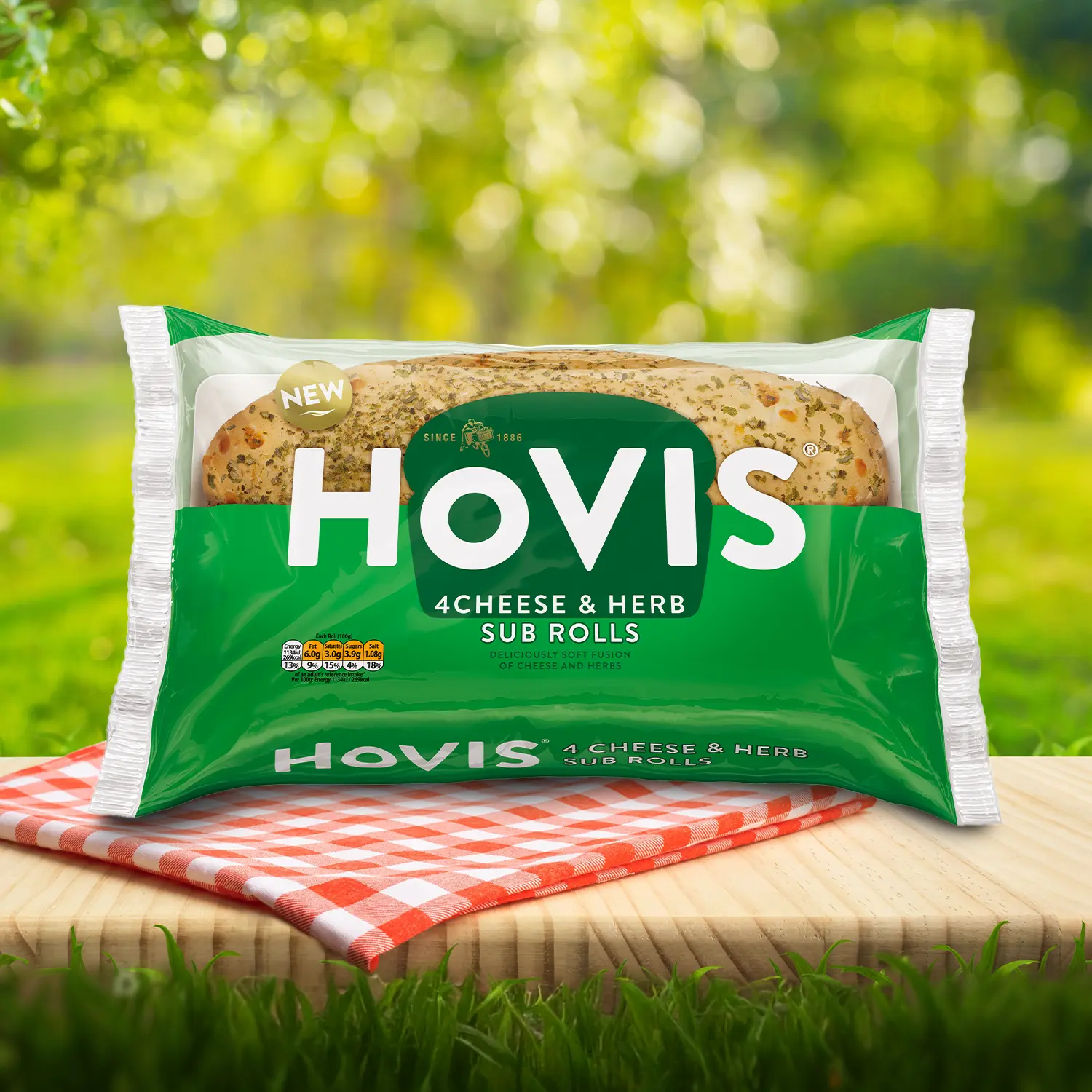 Hovis Cheese & Herb Sub Rolls Sub Rolls packaging on a picnic table with a green outdoor background. The pack shows two soft sub rolls with herbs and highlights “Deliciously soft fusion of cheese and herbs.”  A red gingham cloth adds a picnic feel to the setting.