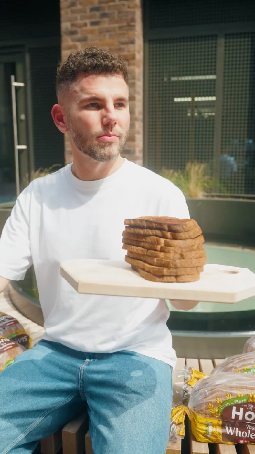 A young man sitting outdoors while holding out a light wooden serving board laden with a high stack of toast.