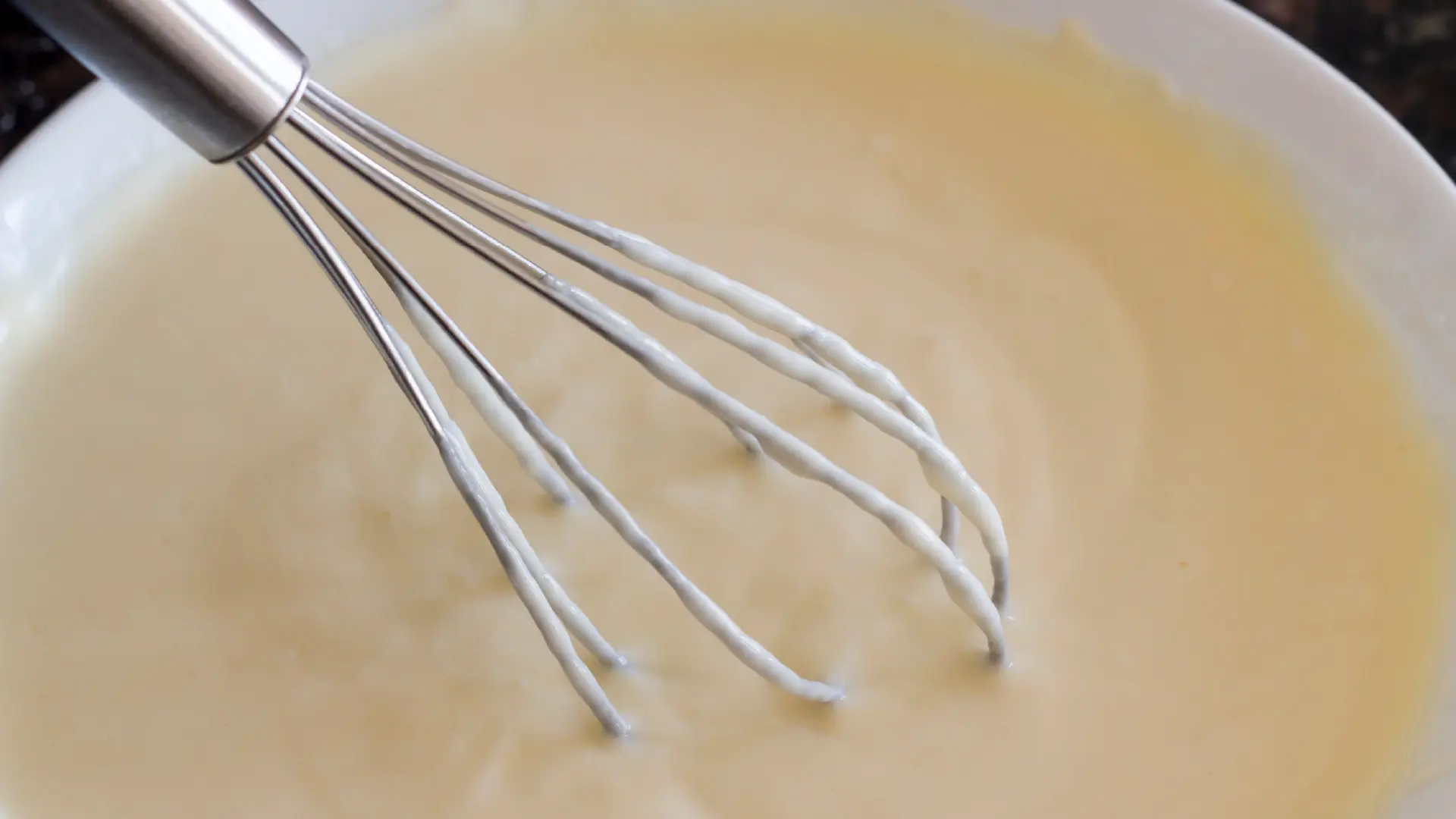 A stainless steel wire whisk rests in a white bowl filled with smooth, creamy light-yellow custard mixture.