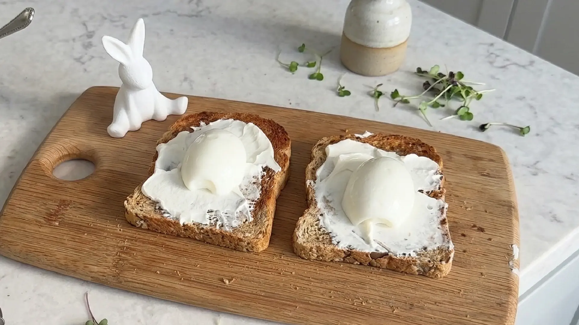 Two slices of seeded toast spread with cream cheese, each topped with a peeled hard-boiled egg, on a wooden cutting board.