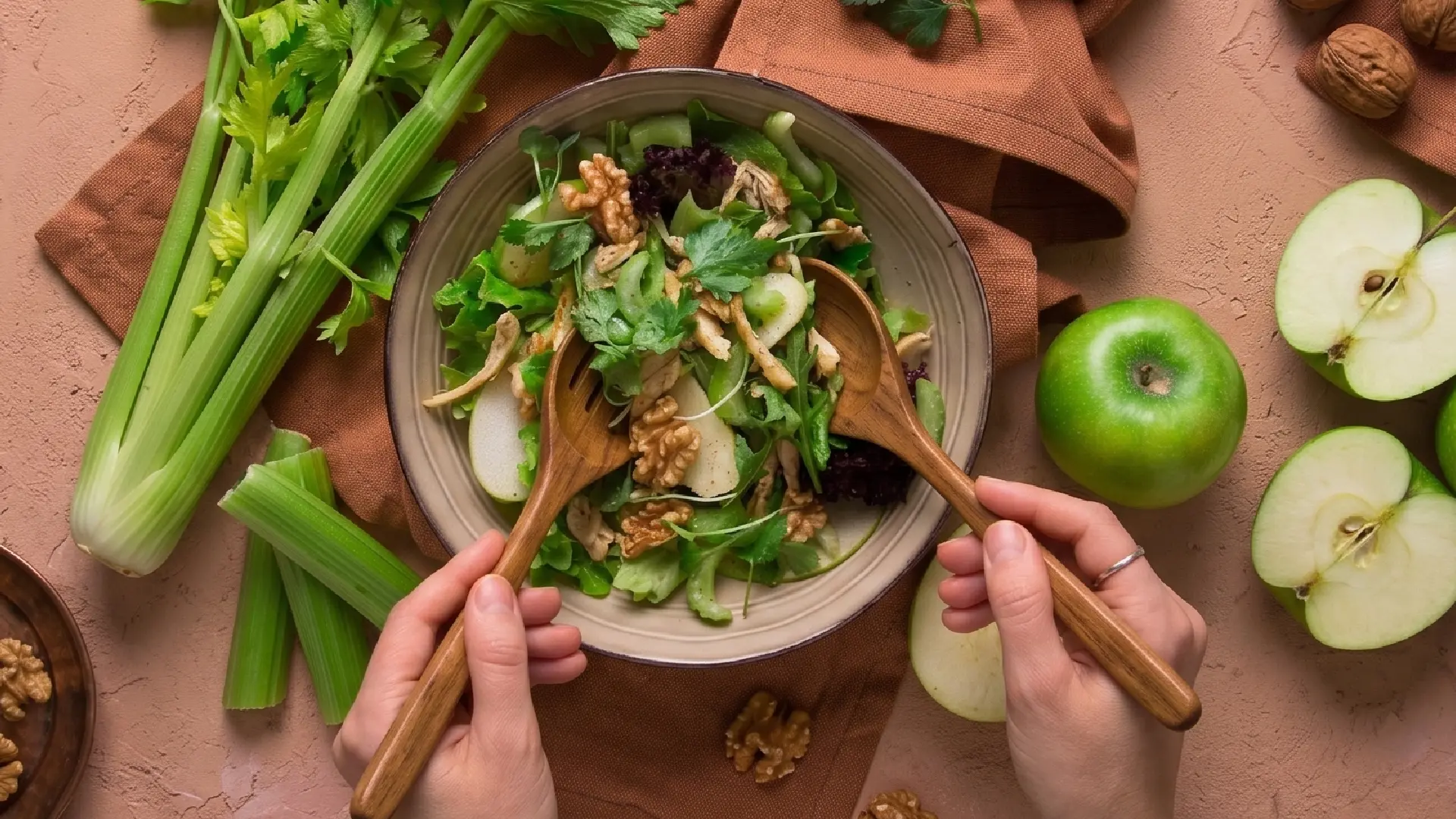 Top-down view of hands tossing a Waldorf salad with wooden spoons, surrounded by fresh celery, walnuts and green apples.