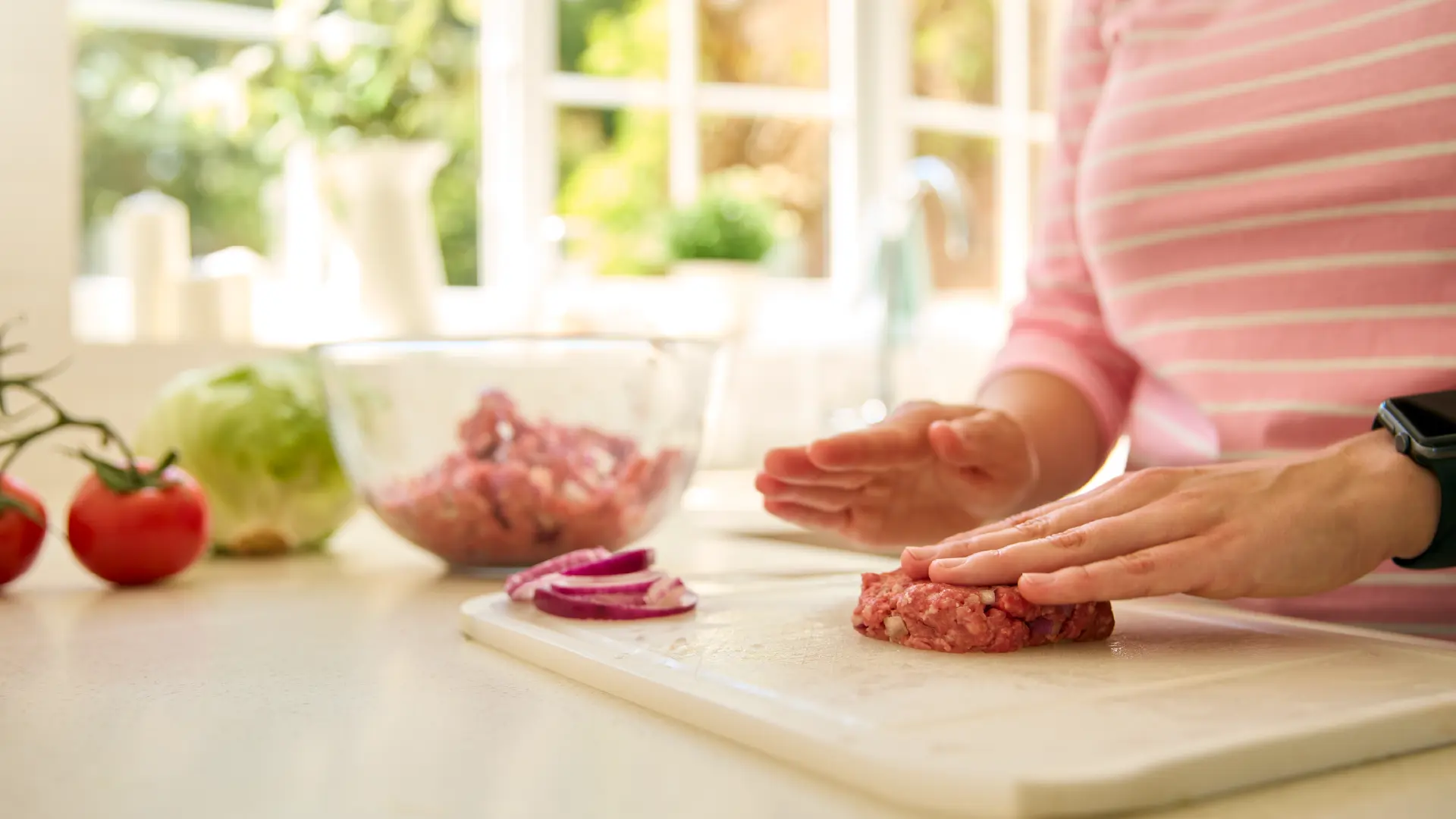 A person wearing a pink striped shirt shapes ground meat into a burger patty on a white cutting board in a bright kitchen.